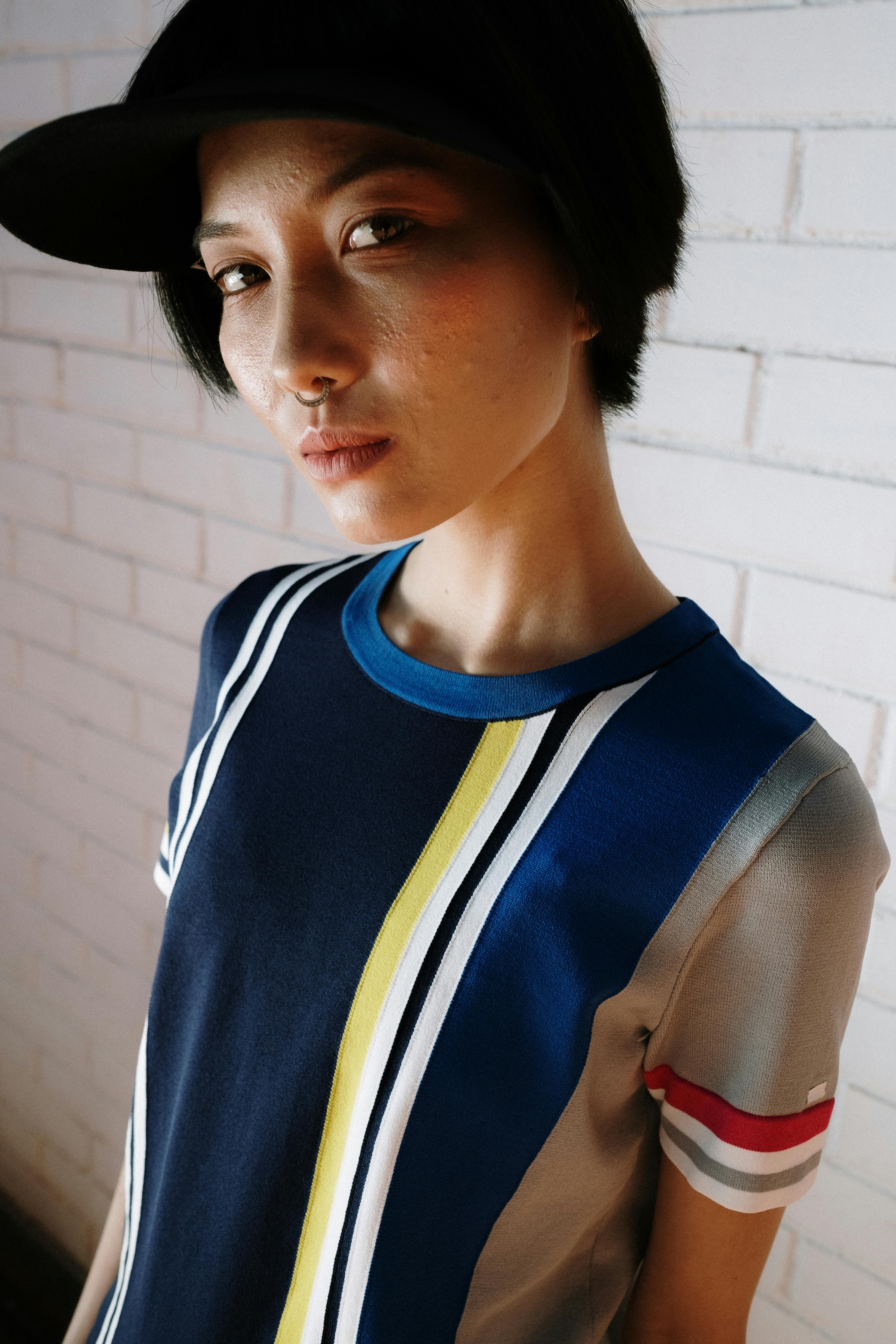 Asian woman with septum piercing wearing a baseball cap and striped shirt against a white brick wall.