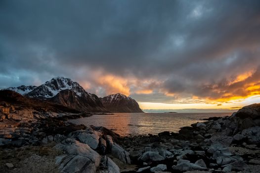 Picturesque scenery of cloudy sunset sky above waving ocean with rocky coast near snowy mountains in Norway