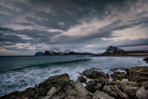 Powerful ocean with foamy waves splashing on rocky beach near snowy mountains against overcast sky in Norway