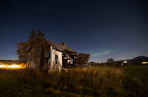 Old ruined hut located on grassy field in countryside against night starry sky with northern light in Norway