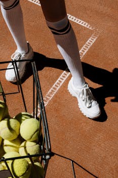 Close-up of tennis shoes and ball basket on a clay court, ready for play.