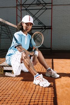A stylish female tennis player sitting on a clay court, holding a racket, ready for action.