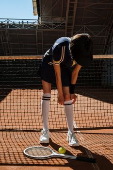 Tennis player adjusting gear on a clay court, with racket and ball on the ground.