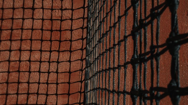 Detailed close-up of a tennis net casting shadows on a clay court surface during the day.