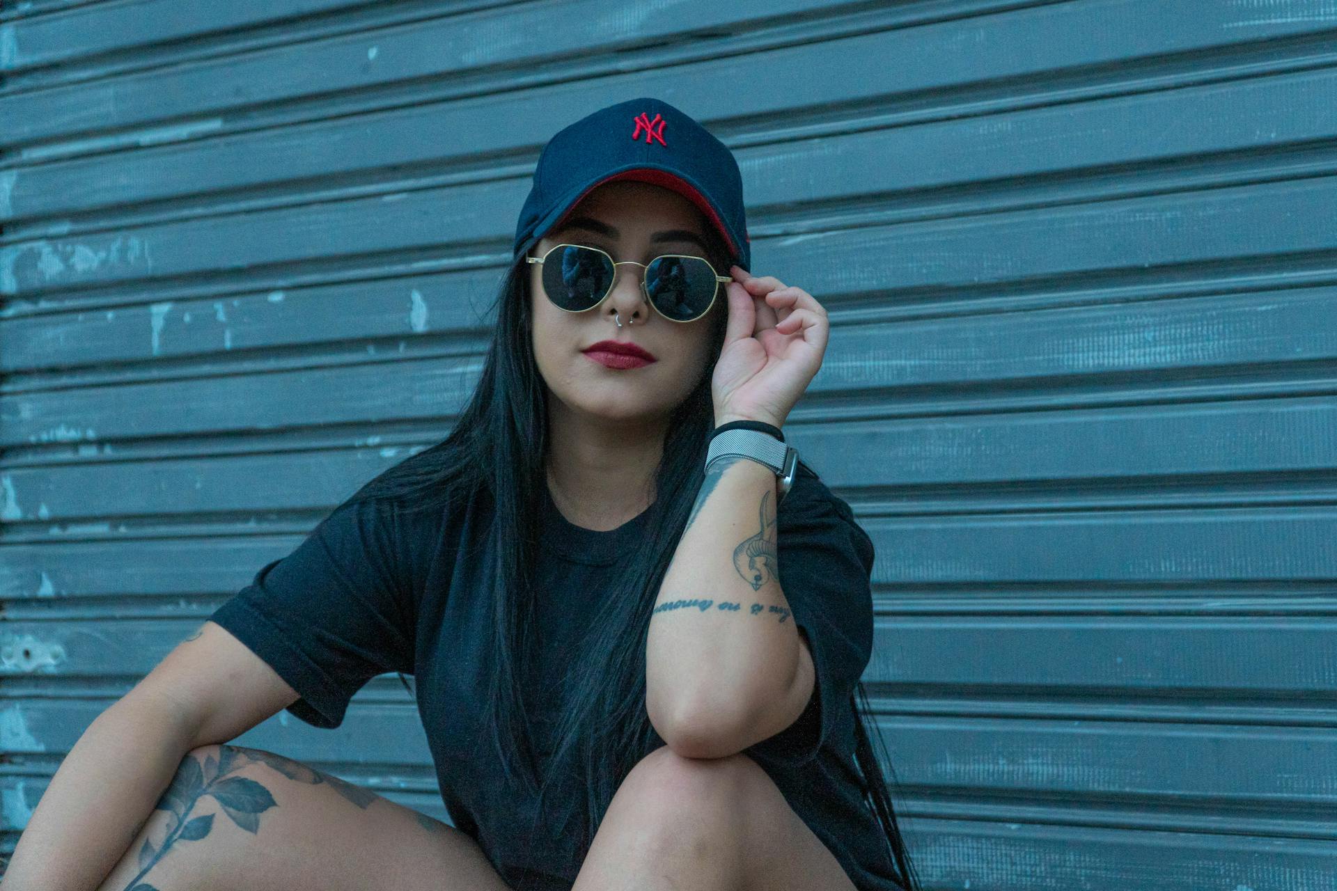 Stylish woman with tattoos in a cap and sunglasses sitting against a metal background.