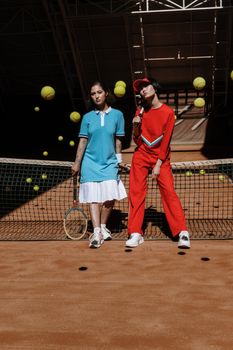 Two women pose confidently on a tennis court with sportswear, surrounded by tennis balls.