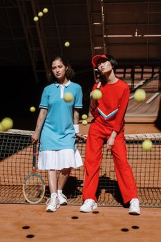 Two women pose on a tennis court amidst bouncing balls, showcasing stylish sportswear.
