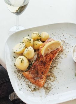 Close-up of baked salmon with potatoes, herbs, and lemon slice on a ceramic plate.