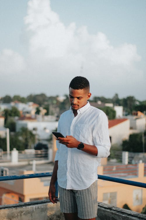 Young African American male in stylish clothes standing on rooftop and using mobile phone on sunny day