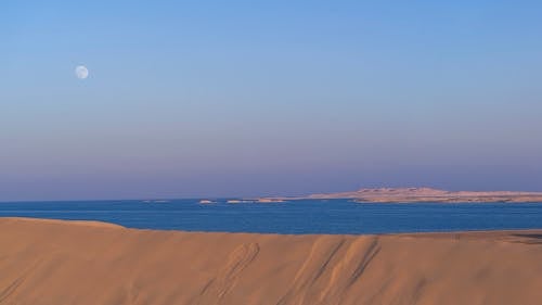 Amazing scenery of blue waving sea surrounded by sands of desert against cloudless sunset sky