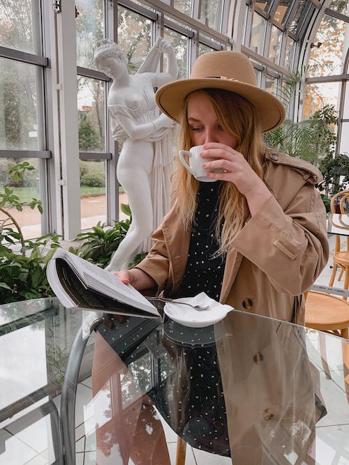 Free Concentrated young female with blond hair in trendy outfit and hat reading magazine while having coffee break in elegant cafe Stock Photo