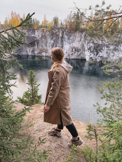 Side view of unrecognizable young female traveler in stylish outfit walking on rocky hill with growing trees and admiring rippling lake against cloudy sky