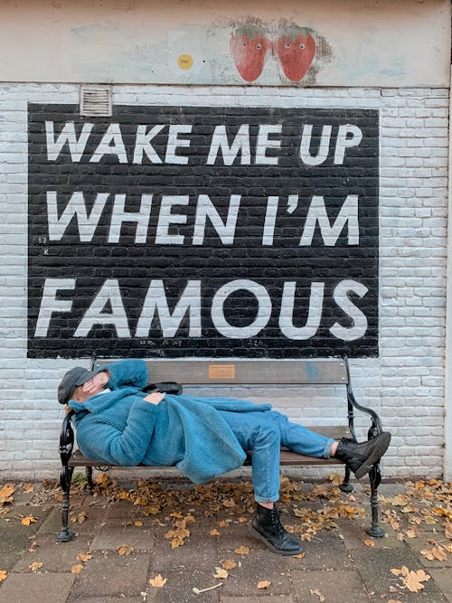 Side view of anonymous lady in fashionable coat and jeans covering face with hand while lying on bench placed on street near Wake Me Up When I M Famous inscription