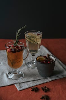 A warm, inviting setup of three mulled wines garnished with cranberries, rosemary, and spices on a fabric backdrop.