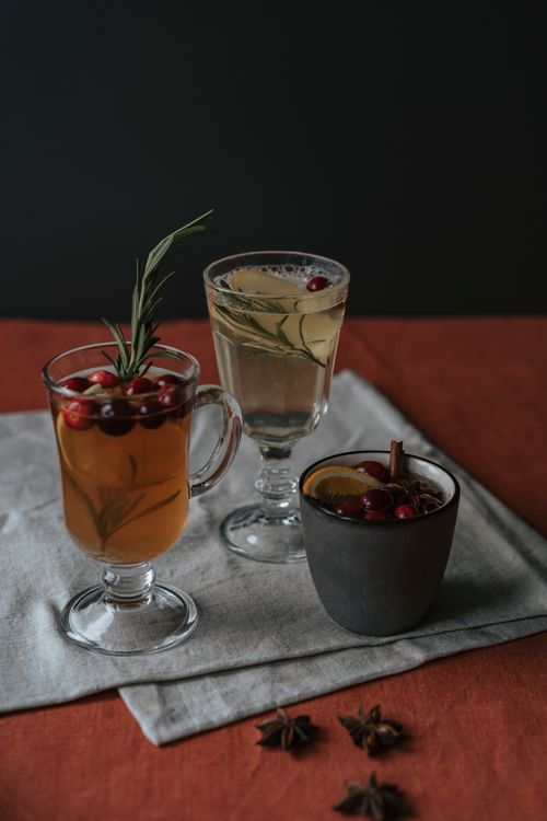 Free A warm, inviting setup of three mulled wines garnished with cranberries, rosemary, and spices on a fabric backdrop. Stock Photo