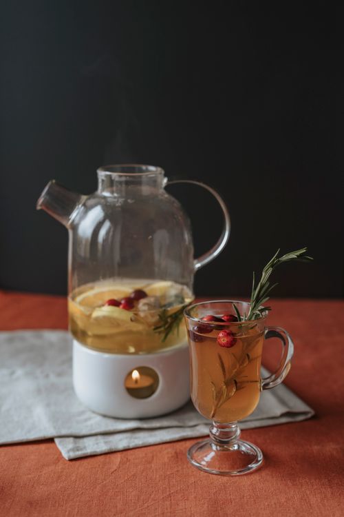 Free Glass cup of herbal tea with lemon slices and cranberries, garnished with rosemary, against a dark background. Stock Photo