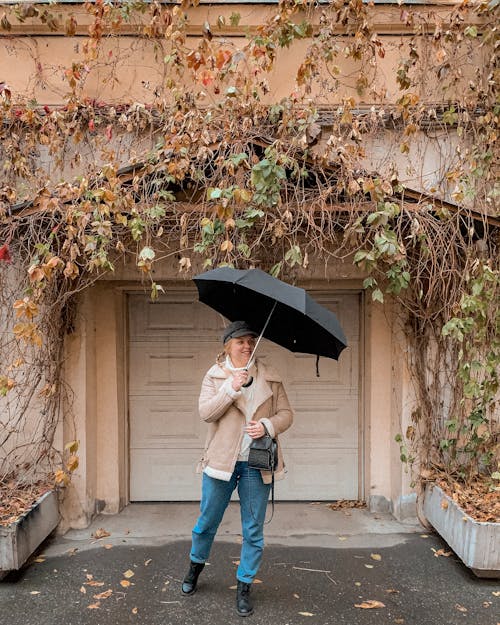 Full length of positive young female tourist in stylish outerwear and hat standing near residential building covered with autumn tree branches with umbrella and looking away
