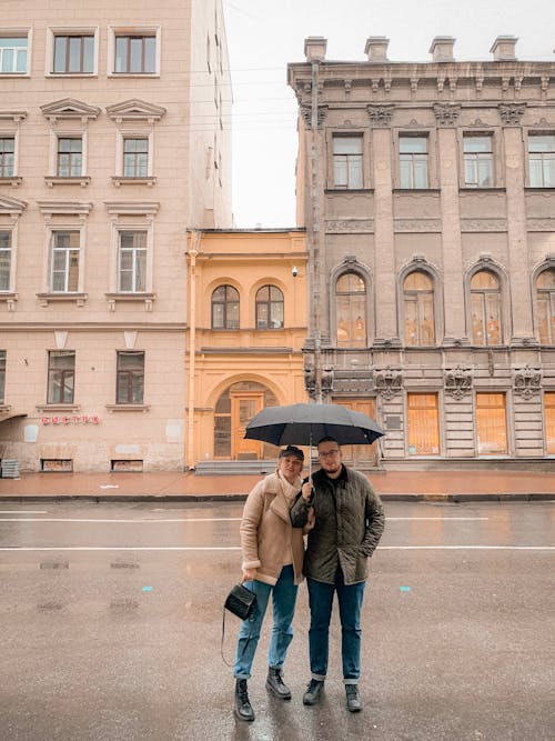 Full body of young stylish man and woman in warm clothes relaxing on street with umbrella near aged buildings during sightseeing tour in city on rainy day