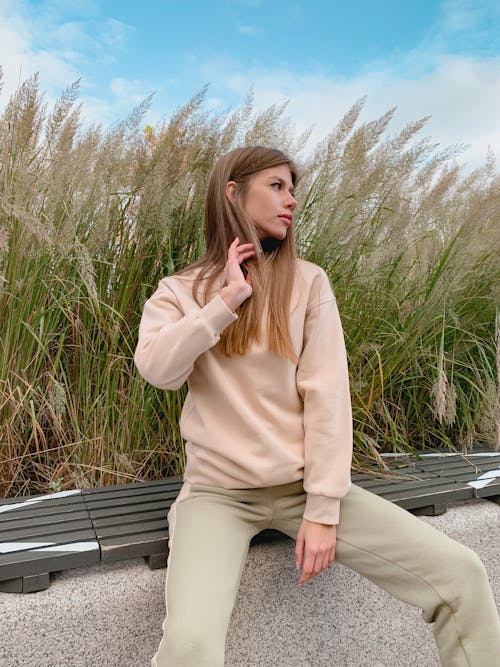 Confident young female in trendy outfit touching long hair and looking away while relaxing on bench in park on sunny day