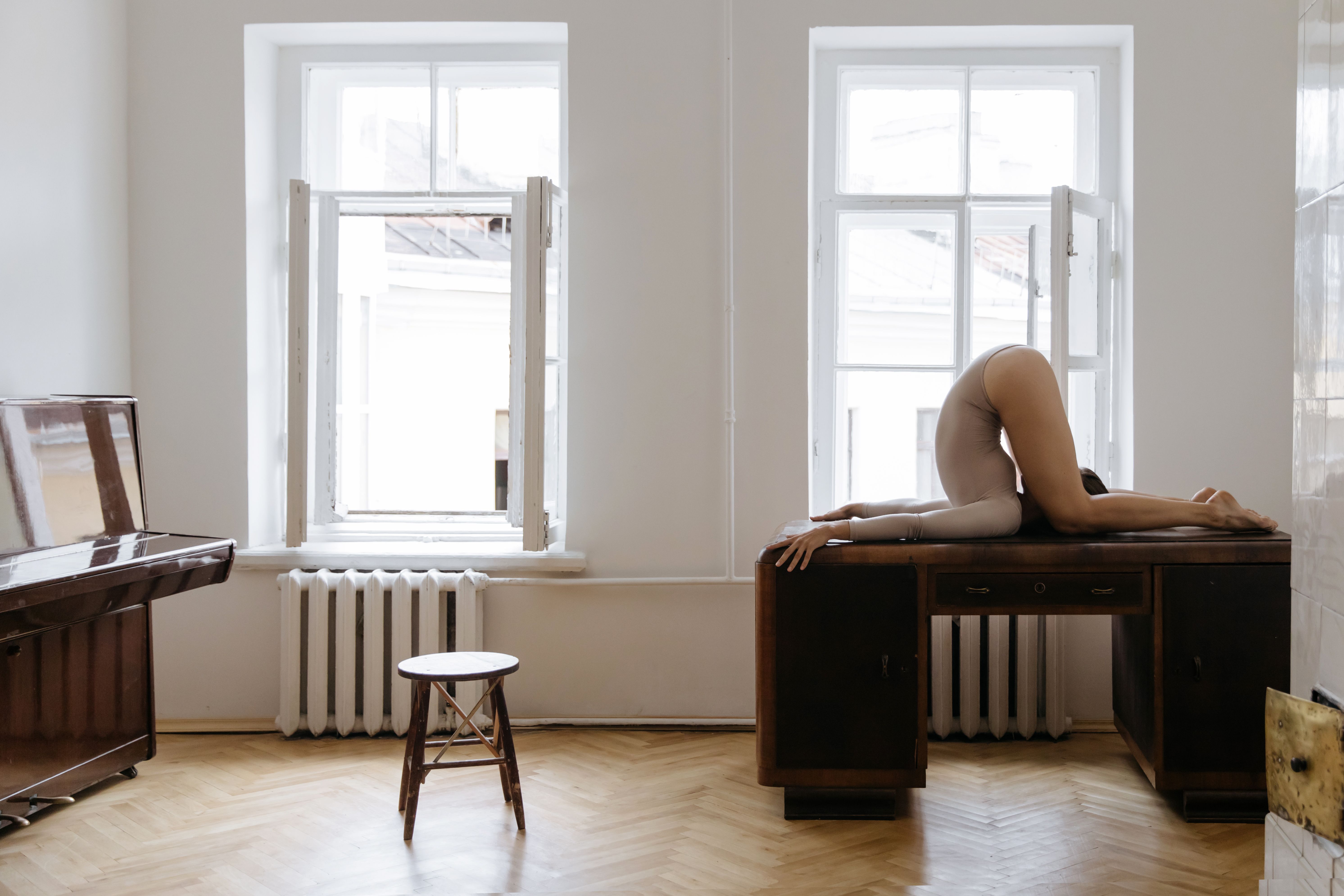 Artistic indoor photo of an adult posing acrobatically on a wooden desk between two windows.