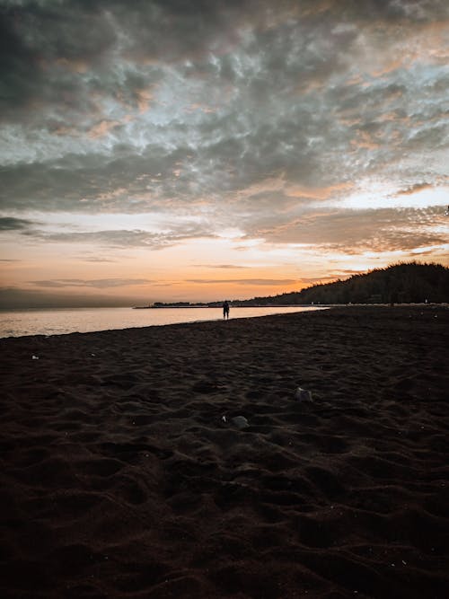 Silhouette of distant unrecognizable traveler walking on sandy seashore along calm sea against cloudy sundown sky on evening time in nature