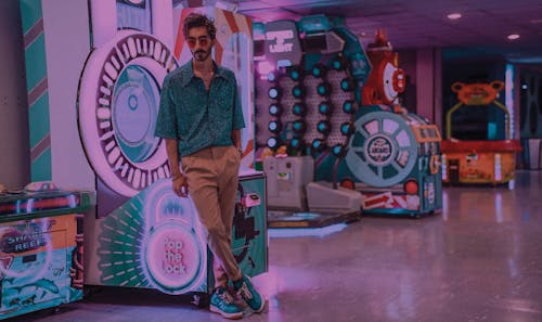 Full body of thoughtful bearded male wearing sunglasses standing with legs crossed and hand in pocket near slot machines in shopping center in city