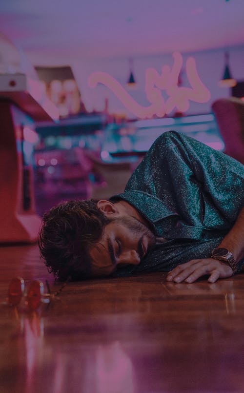 Bearded male with sunglasses wearing shirt lying on floor in shopping center at low light on blurred background