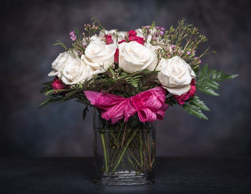 Free Elegant bouquet with fresh white and pink roses with fern leaves and wildflowers decorated with ribbon and placed in glass vase against black background Stock Photo