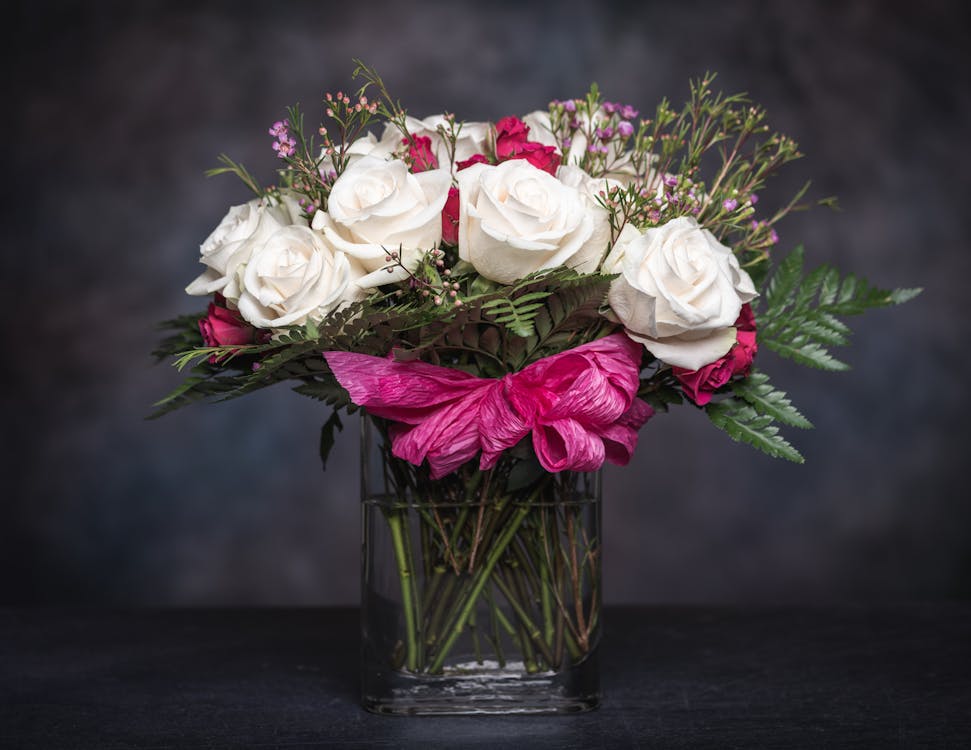 Elegant bouquet with fresh white and pink roses with fern leaves and wildflowers decorated with ribbon and placed in glass vase against black background