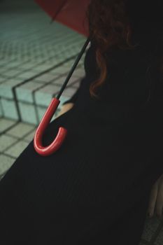 一位长卷发的匿名女子，身穿黑色外套，带着红色雨伞，坐在瓷砖楼梯上