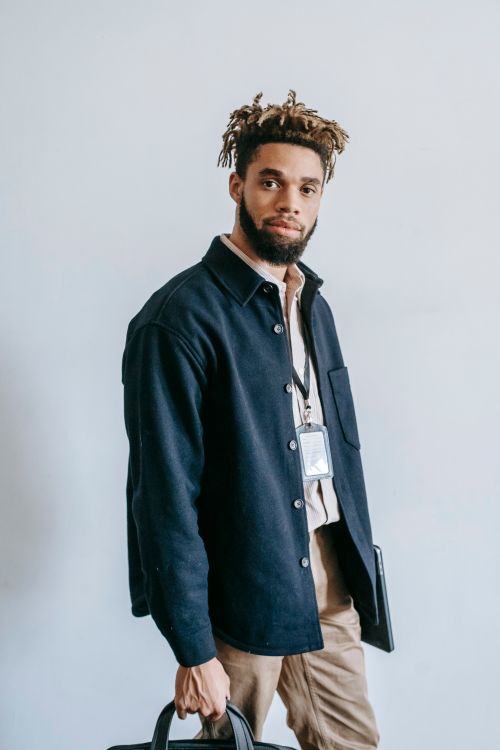 Free Confident young African American male in trendy outfit with cardholder on neck carrying briefcase while standing against white wall in studio and looking at camera Stock Photo