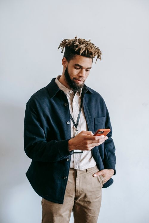 Free Content young African American male with dreadlocks wearing trendy clothes standing with hand in pocket and browsing modern mobile phone in light studio Stock Photo