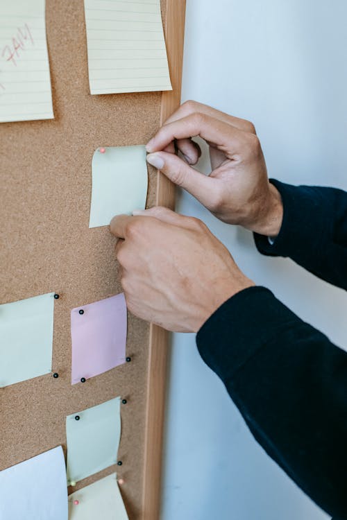 Crop unrecognizable male attaching colorful memo papers with thumbtacks to cork bulletin board hanging on white wall in office