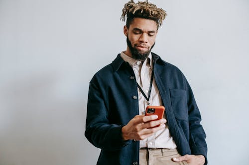Self assured young bearded African American male employee browsing mobile phone while standing against white background with hand in pocket
