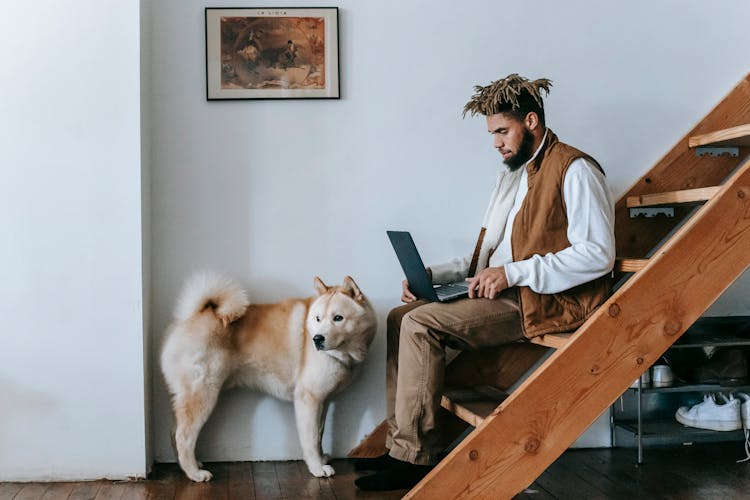 Young Black Man Working Online On Laptop Sitting On Stairs Near Dog At Home