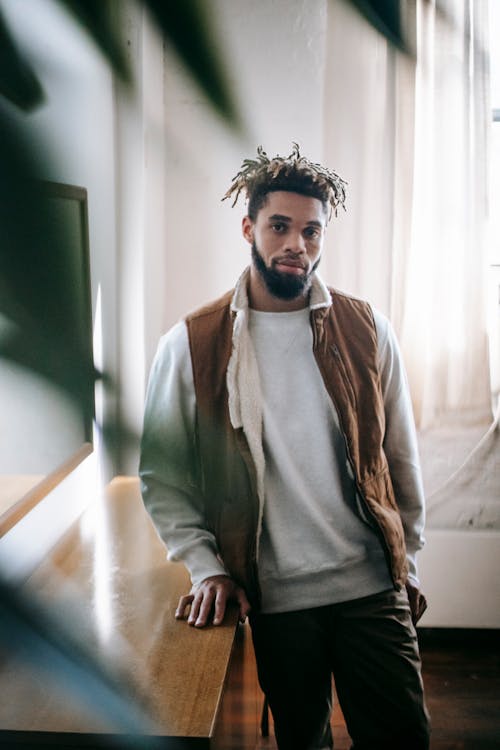 Thoughtful young bearded ethnic male millennial with dreadlocks in stylish outfit standing near wooden table in modern apartment and looking at camera