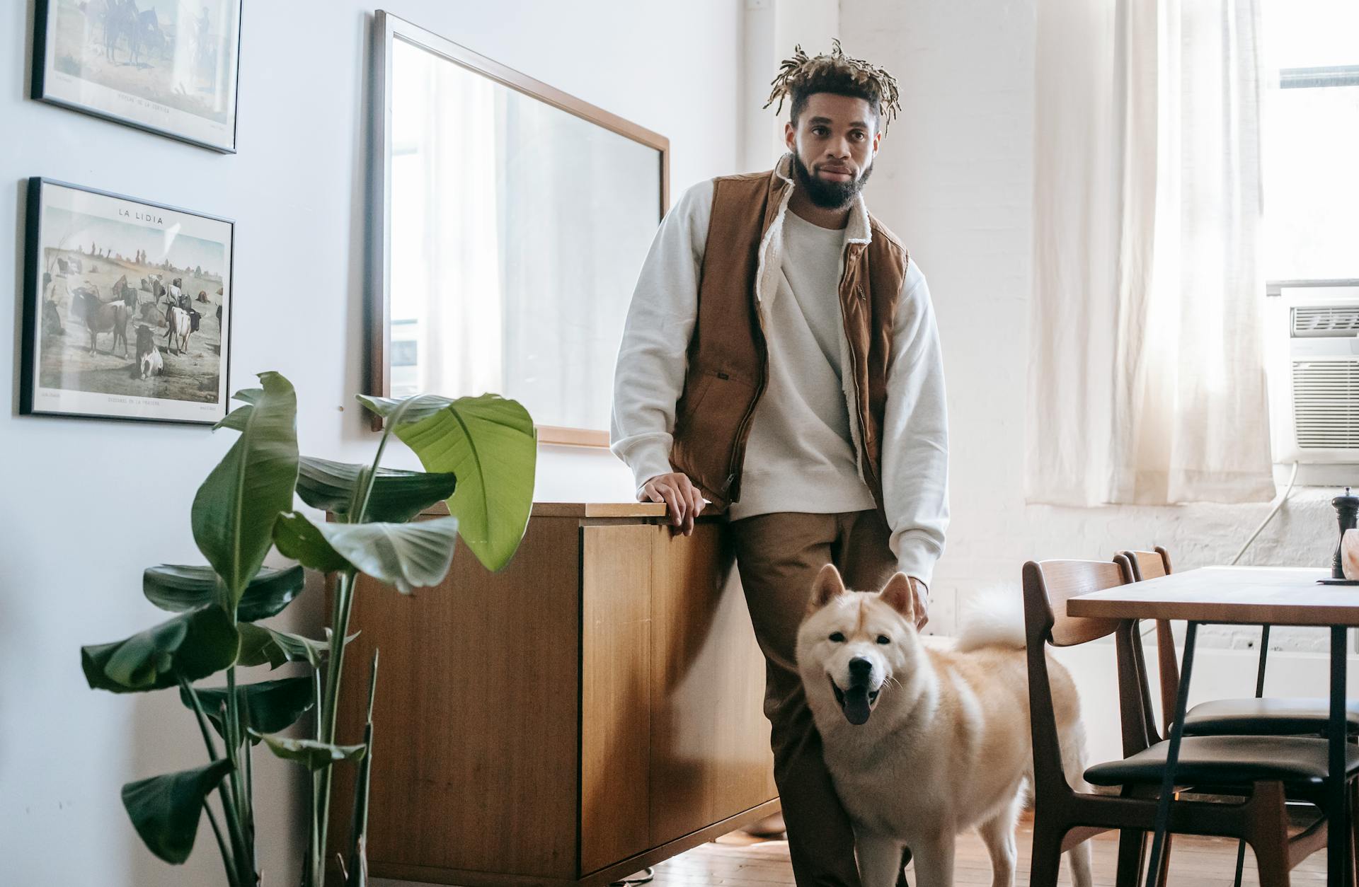 En lugn ung afroamerikansk manlig ägare med dreadlocks och skägg står i ett mysigt kök med en söt Akita Inu-hund