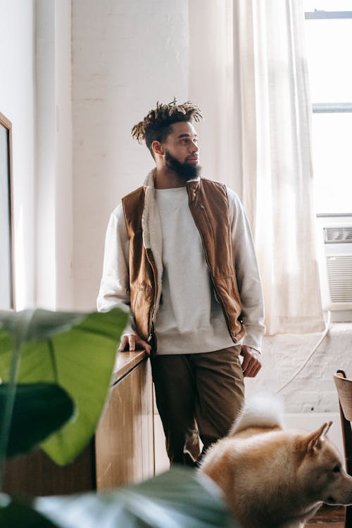Dreamy young bearded African American male owner in stylish clothes standing in light apartment near cute Akita dog and looking away
