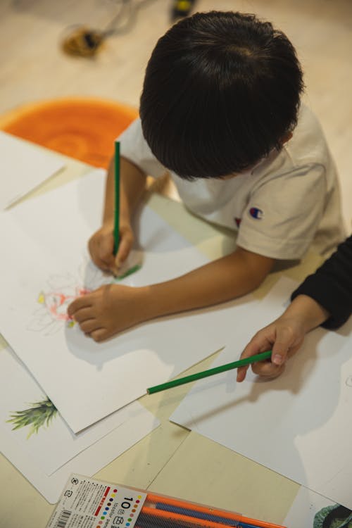 High angle of crop anonymous ethnic children drawing on paper sheet with pencils during art class in kindergarten