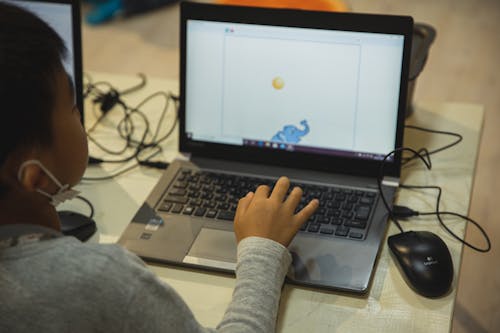 From above back view of crop anonymous Asian schoolchild typing on netbook with illustration on screen during lesson in school