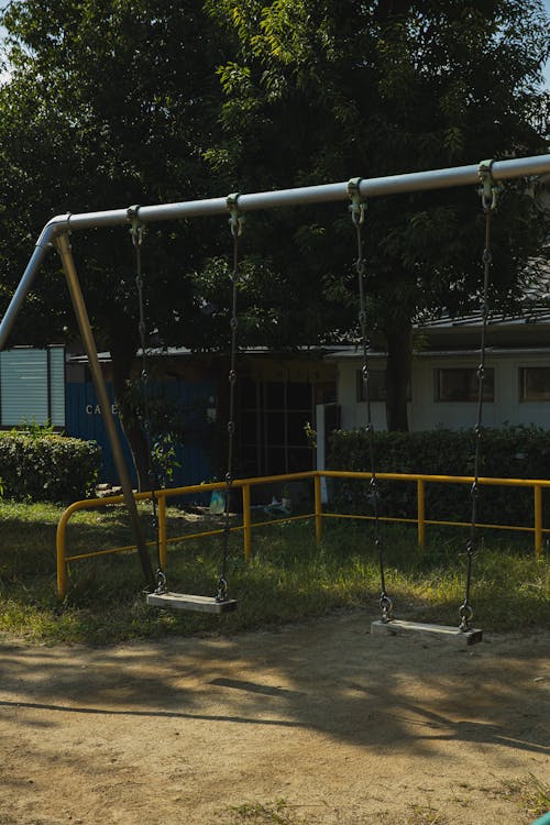 Metal empty swings on playground in yard among green trees on sunny summer day