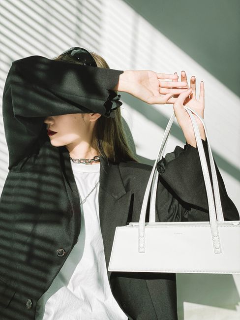 Young female model covering face wearing trendy oversize black jacket and t shirt while holding modern handbag