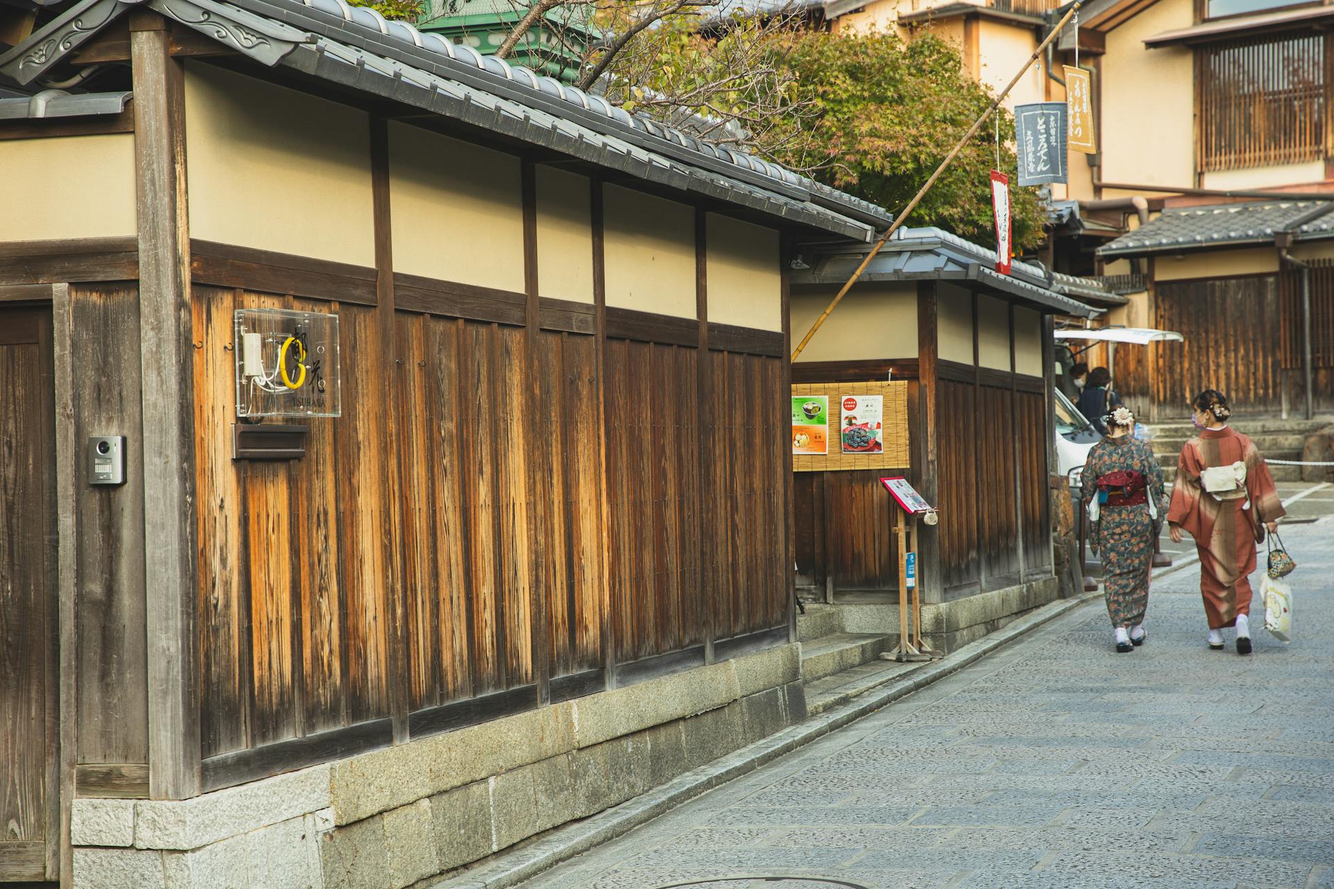 Back view of unrecognizable local Japanese ladies in traditional kimonos walking on paved street near typical aged wooden houses in Kyoto