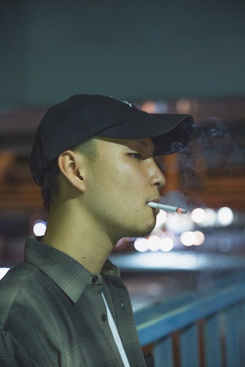 Side view of Asian male smoker with cigarette standing near fence at night and looking away