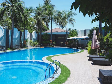 Outdoor swimming pool with sunbeds on territory of tropical hotel with green palm trees against cloudless blue sky