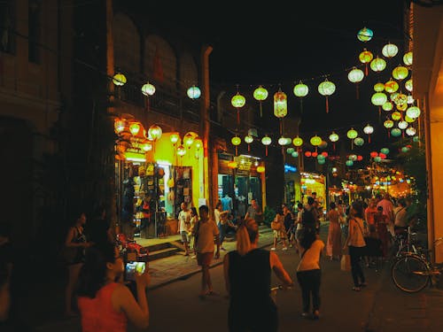 Crowd of unrecognizable diverse tourists walking on narrow pedestrian street with aged houses and cafes in city old district decorated with Asian paper lanterns at night