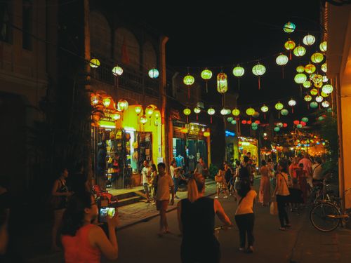Free Crowd of unrecognizable diverse tourists walking on narrow pedestrian street with aged houses and cafes in city old district decorated with Asian paper lanterns at night Stock Photo