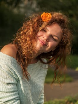 A young woman with curly hair and a flower accessory smiling in a sunny outdoor setting.