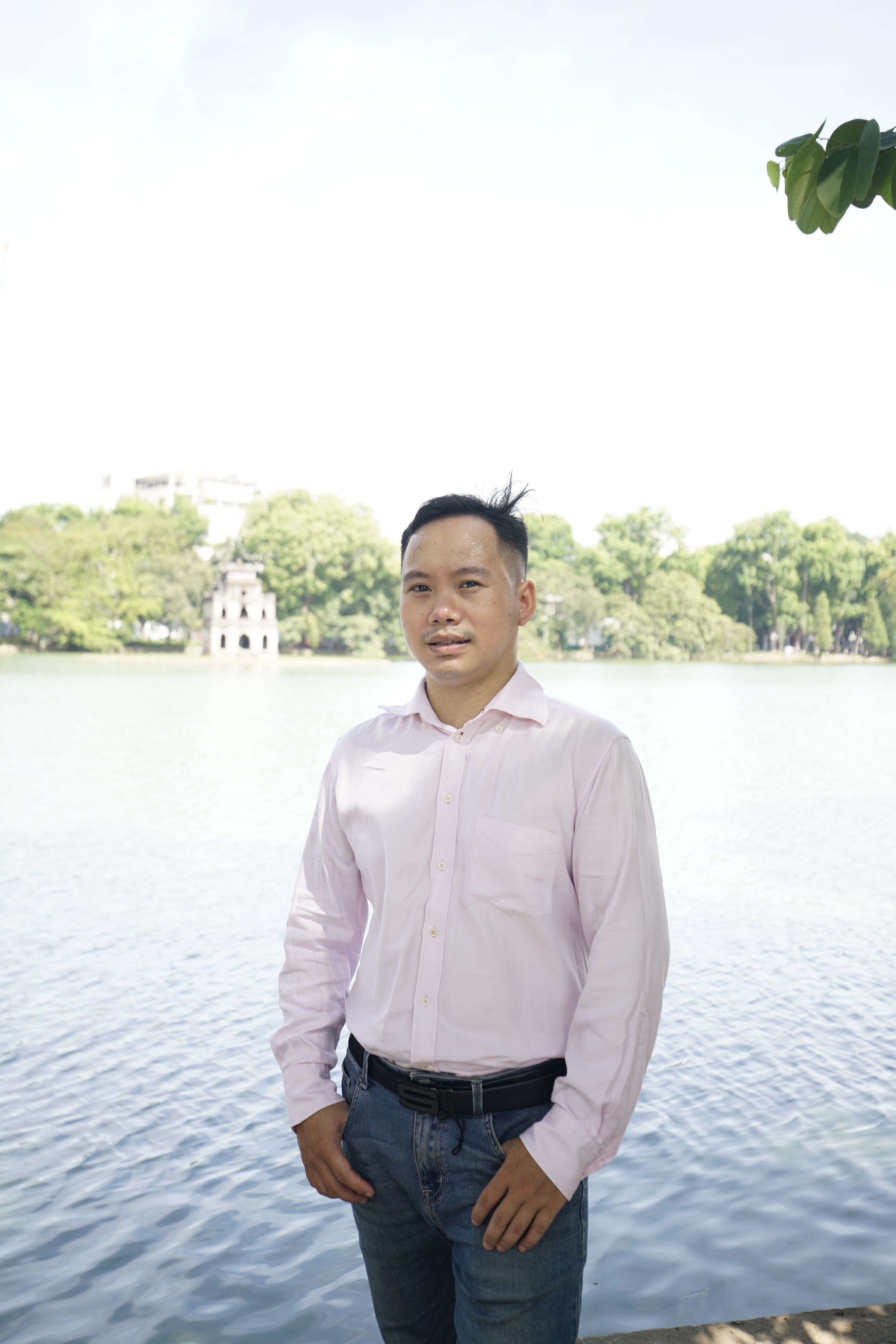 Young Asian man in a long sleeve shirt standing by a serene river under a clear sky.