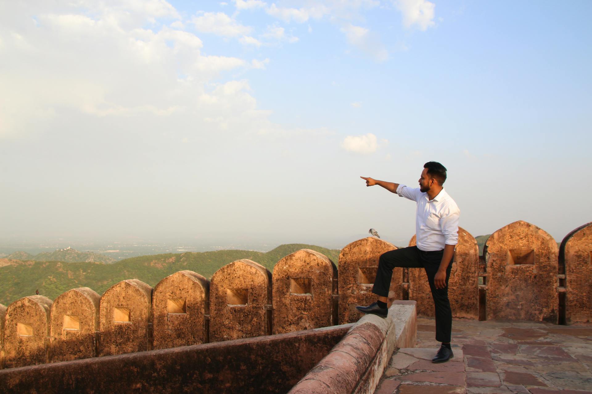 A man in formal attire stands on a historic fort wall, pointing towards a scenic distant horizon.